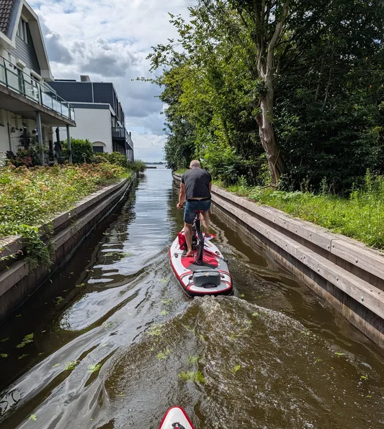 Man vaart op een Red Shark door een smalle sloot met houten beschoeiing tussen Nederlandse huizen en bomen, met een tweede board erachteraan